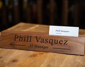 Puede incluir: Una placa de nombre de escritorio de madera con el nombre "Phill Vasquez" y el título "IT Manager" grabado en ella. Una tarjeta de visita con el mismo nombre y título se muestra en una ranura en la placa de nombre.