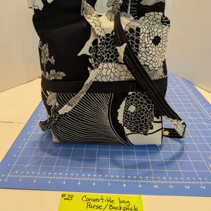 May include: A black and white fabric bag featuring a floral pattern with two shoulder straps. A yellow note in front displays the text #23 Convertible bag Purse/Backpack, Colors Black & White, Size approx 23x36 cm, placed on a blue cutting mat.