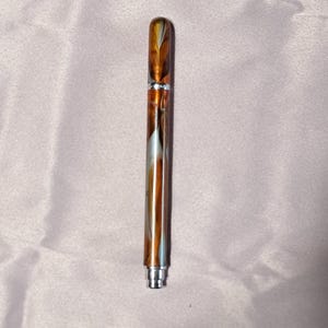 May include: A cylindrical pen featuring a marbled pattern of amber, brown, and white swirls. The writing instrument has a polished silver-colored metal band separating the cap from the body, with a matching silver-colored tip at the bottom end.