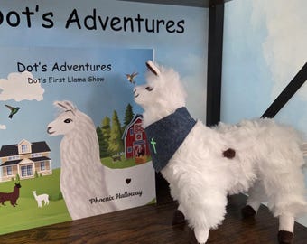 Op de afbeelding: Een wit pluche lama-speeltje met een blauwe bandana met een groen kruis, naast een kinderboek getiteld Dot's Adventures: Dot's First Llama Show door Phoenix Halloway, met een illustratie van een lama, een huis en een schuur.