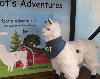 Op de afbeelding: Een witte pluche lama met een blauwe bandana met de letters DOT in geel en groen, naast een kinderboek getiteld Dot's Adventures: Dot Moves to a New Home door Phoenix Halloway, met een illustratie van een lama op de kaft.
