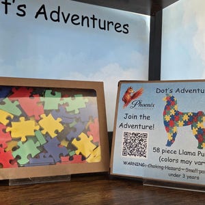 May include: A 58-piece llama puzzle in a brown box with a clear window showing colorful puzzle pieces. Next to it, a display card features the text Dot's Adventures, a llama graphic, a QR code, and the finished size of 13 inches by 9 inches.