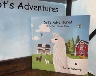 Op de afbeelding: Een vierkant kinderboek getiteld Dot's Adventures: Dot's First Llama Show door Phoenix Halloway. De kaft toont een witte lama op de voorgrond, een rode schuur, een huis, groene velden en kolibries tegen een lichtblauwe lucht als achtergrond.