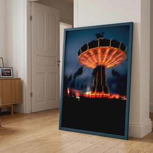 May include: A framed wall art print featuring a long-exposure photograph of a spinning amusement park swing ride at night. The ride is illuminated with bright orange and yellow lights, creating a motion blur effect against a dark blue sky.