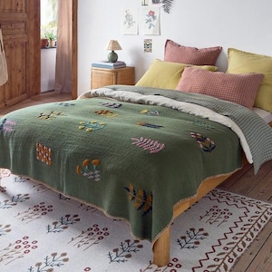 Puede incluir: Una cama con una colcha texturizada de color verde salvia bordada con motivos florales y botánicos coloridos. La ropa de cama incluye dos almohadas cuadradas de color amarillo mostaza, una almohada rectangular de cuadros rojos y un edredón de cuadros verde claro.