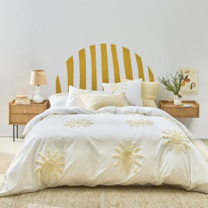 Puede incluir: Una funda nórdica blanca con un patrón repetitivo de soles amarillos con relieve, acompañada de almohadas blancas y amarillas a juego. La cama se sitúa frente a una pared con un gran diseño de arco solar amarillo pintado y dos mesitas de noche de ratán.
