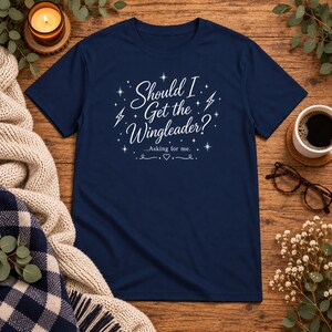 Peut inclure: Un t-shirt bleu marine à manches courtes avec le texte blanc "Should I Get the Wingleader? ...Asking for me." Le motif est agrémenté d'étoiles graphiques blanches, d'éclairs et d'un petit cœur centré sous le texte imprimé.