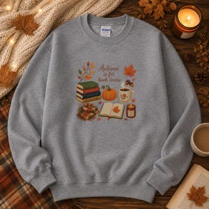 Puede incluir: Una sudadera gris de cuello redondo con un estampado gráfico que incluye el texto Autumn is for book lovers. El diseño muestra una pila de libros, una calabaza, una taza de café, un libro abierto con una hoja, una bufanda a cuadros y bellotas.
