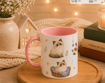 Może przedstawiać: Biały ceramiczny kubek z różowym wnętrzem i uchwytem, ozdobiony ilustracjami puszystych kociąt, małych kwiatów i serc. Kubek ma pojemność 325ml i zawiera ikony wskazujące, że można go używać w kuchence mikrofalowej i myć w zmywarce.