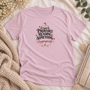 Op de afbeelding: Een lichtroze T-shirt met korte mouwen en een grafisch ontwerp met een open boek en de tekst "Probably Reading Something Inappropriate" in zwarte en rode letters, omringd door kleine rode hartjes en decoratieve gouden sterren.