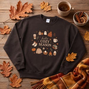 Peut inclure: Un sweat-shirt à col rond marron foncé avec un motif central portant l'inscription "hello COZY SEASON" entouré d'illustrations de bougie, fantôme, bonnet, tasses de café, chaussettes, citrouille, pull, cupcake, feu de camp et livres.