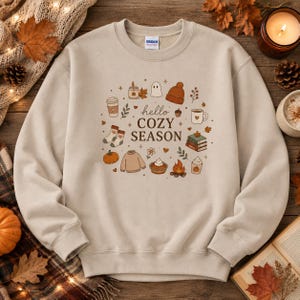 Pode incluir: Uma camisola bege de gola redonda com um gráfico central com o texto "hello COZY SEASON" rodeado por ilustrações de uma abóbora, meias, uma camisola, uma tarte, uma fogueira, um fantasma, um gorro, uma chávena e livros.