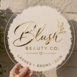 May include: A circular clear acrylic sign featuring a textured cream-coloured background. The sign displays the gold-coloured text "Blush BEAUTY CO." with a small heart icon and the words "LASHES • BROWS • SKIN" below, accompanied by a gold leaf graphic.