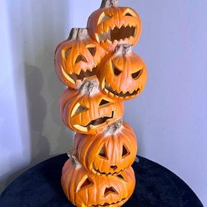 Può includere: Una pila verticale di cinque zucche di Halloween arancioni con occhi triangolari intagliati e bocche frastagliate. Ogni zucca presenta una superficie strutturata con steli marroni e creste profonde, creando una scultura decorativa alta e singola.