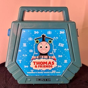 Może przedstawiać: Morska plastikowa walizka z grafiką Thomas & Friends z przodu. Walizka posiada uchwyt i zawiera napis Thomas & Friends, Collector's Really Useful Carry Case oraz ERTL na niebieskim tle z białymi ikonami przedstawiającymi różne przedmioty.