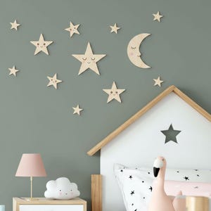 Può includere: Un set di adesivi murali in legno chiaro raffiguranti una luna crescente e varie stelle di diverse dimensioni. Diverse stelle e la luna presentano volti stampati con occhi chiusi e guance rosa, disposti su una parete.