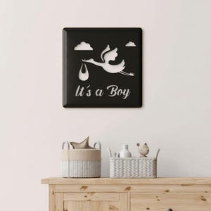 May include: A square black wall sign featuring a white cutout silhouette of a stork carrying a bundle. Above the stork are two small white clouds. Below the stork, the text It's a Boy is written in a white cursive script font.