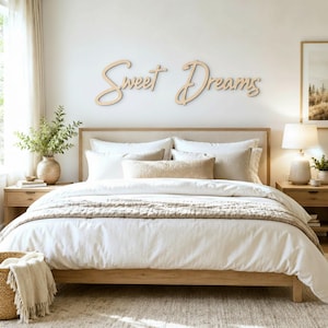 May include: A light-colored wooden wall sign featuring the cursive text Sweet Dreams mounted above a bed. The sign consists of two separate script words made from a smooth, natural wood material, displayed horizontally against a plain wall.