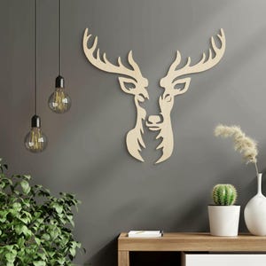 Op de afbeelding: Een lichtgekleurd houten wanddecoratie-item met een gestileerd, symmetrisch silhouet van een hertenkop met groot, vertakt gewei. Het ontwerp bestaat uit twee gespiegelde helften die in het midden samenkomen, wat zorgt voor een vlakke uitstraling.