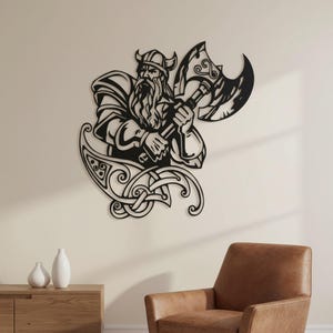 Op de afbeelding: Een zwart metalen wanddecoratie met een gestileerde Viking-krijger met een lange baard en een gehoornde helm, die een grote dubbelbladige bijl vasthoudt. Het ontwerp bevat ingewikkelde, krullende knooppatronen aan de basis van de figuur.