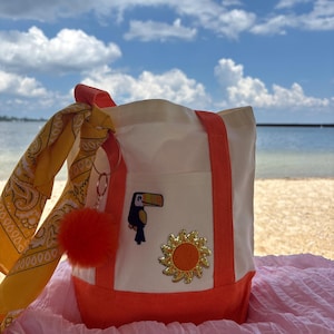 May include: A white canvas tote bag with orange trim, featuring a toucan patch and a sun patch on the front pocket. A yellow paisley bandana is tied to the handle, accompanied by a round orange pom-pom charm attached to a metal ring.