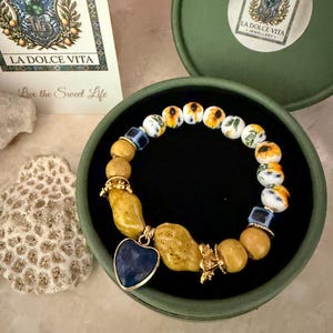 Puede incluir: Una pulsera de cuentas con cuentas de cerámica blanca con patrones de girasoles amarillos, cuentas texturizadas color mostaza, pequeñas cuentas cuadradas azules y un colgante azul oscuro en forma de corazón con detalles de metal dorado.