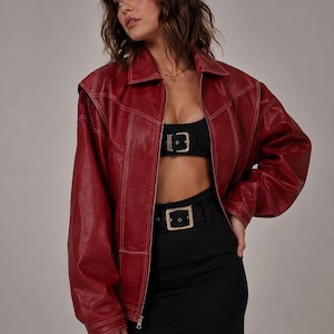 May include: A deep red oversized jacket with a pointed collar, long sleeves, and prominent white contrast stitching along the seams. The jacket features a front zipper closure and is worn over a black cropped top and a black high-waisted skirt with a belt.