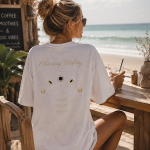 May include: A white short-sleeved t-shirt featuring the text Chasing Totality in gold script above a graphic of moon phases. Below the moon phases, the dates 21.08.2017, 08.04.2024, and 12.08.2026 are printed in a simple gold font on the back.