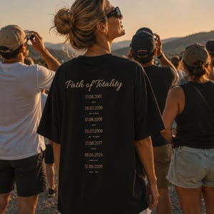Pode incluir: Uma t-shirt preta de manga curta com o texto dourado Path of Totality e uma lista de datas incluindo 21.06.2001, 29.03.2006, 01.08.2008, 22.07.2009, 21.08.2017, 08.04.2024 e 12.08.2026 impressas nas costas da peça.