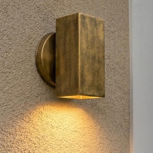 May include: A rectangular wall-mounted light fixture with an aged brass finish. The fixture features a cylindrical base plate attached to the wall and a box-shaped housing that directs light downward, casting a warm glow onto the textured surface.