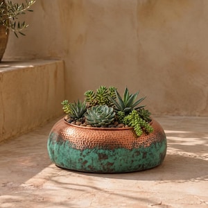 May include: A round, hammered metal planter with a copper-toned upper rim and a weathered, teal-green patina finish on the lower section, filled with a variety of green succulent plants and small dark pebbles arranged in the center.