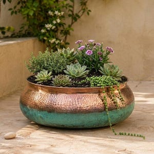 Pode incluir: Um vaso redondo de cobre martelado com acabamento em pátina turquesa na metade inferior. A taça está cheia de terra escura e uma variedade de suculentas verdes, pequenas ervas e um grupo de flores roxas que caem sobre a borda do vaso.