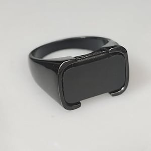 Puede incluir: Un anillo negro sólido que presenta una cara rectangular lisa engastada en una banda pulida. El diseño es monocromático, con un acabado brillante que cubre toda la superficie del anillo, creando un aspecto elegante y uniforme desde cualquier ángulo.