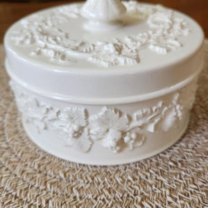 May include: A round, off-white lidded trinket box featuring a raised, molded relief pattern of grapevines and leaves encircling the side and the lid. The lid is topped with a pointed, conical finial handle, all in a smooth, uniform finish.