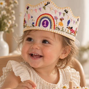 May include: A cream-coloured felt crown featuring intricate embroidery of a central rainbow with the number one, surrounded by colourful bunting, small flowers, a white bird, and a small dog, finished with a yellow stitched scalloped edge.