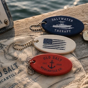 May include: Three oval-shaped keychains with ball chains. The top is dark blue with a white boat graphic and the text SALTWATER THERAPY. The middle is off-white with a distressed American flag. The bottom is red with an anchor and OLD SALT MERCANTILE CO.
