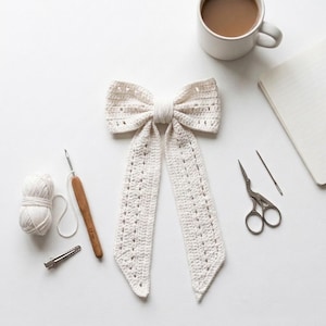 May include: A cream-colored crocheted bow with two long, dangling tails featuring a delicate openwork pattern. The bow is surrounded by crafting supplies including a ball of yarn, a wooden crochet hook, a metal hair clip, and scissors.