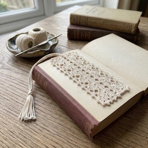 May include: A cream-colored rectangular crochet bookmark featuring an intricate openwork lace pattern and a matching twisted cord tassel. The bookmark is placed inside an open hardcover book, with two additional books and yarn nearby.