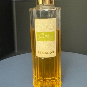 Könnte beinhalten: Eine hohe, zylindrische Glasparfümflasche gefüllt mit bernsteinfarbener Flüssigkeit. Sie hat einen goldfarbenen Verschluss und ein rechteckiges Etikett mit einem hellgrünen Mittelstreifen. Das Etikett zeigt den Text Sortilège, Parfum de Toilette und LE GALION.