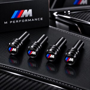 May include: Four black cylindrical valve stem caps featuring the M Performance logo with blue, purple, and red stripes. Each cap has a circular top with the M emblem and a grooved black metal body, presented alongside a black M Performance box.