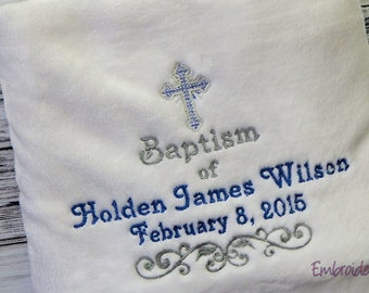 May include: A white, plush blanket embroidered with a cross and the words "Baptism of Holden James Wilson February 8, 2015." The blanket features blue and silver thread, with a decorative scroll design at the bottom. The blanket is a keepsake for a baptism.