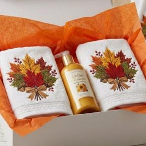 May include: A white gift box lined with orange tissue paper containing two white hand towels and a bottle of lotion. Each towel features an embroidered bouquet of autumn leaves, berries, and a bow. The lotion bottle is labeled Golden Autumn.