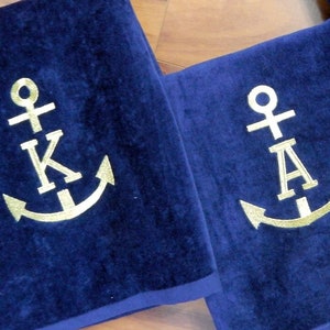 May include: Navy blue bath towels with gold anchor and initial embroidery. The towels are folded and arranged on a wooden surface. The initials are K, A, and S. The towels have a dark blue border.