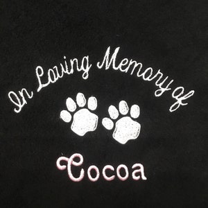 May include: Black fabric embroidered with the words "In Loving Memory of" above two white paw prints. Below the paw prints, the name "Cocoa" is embroidered in pink.