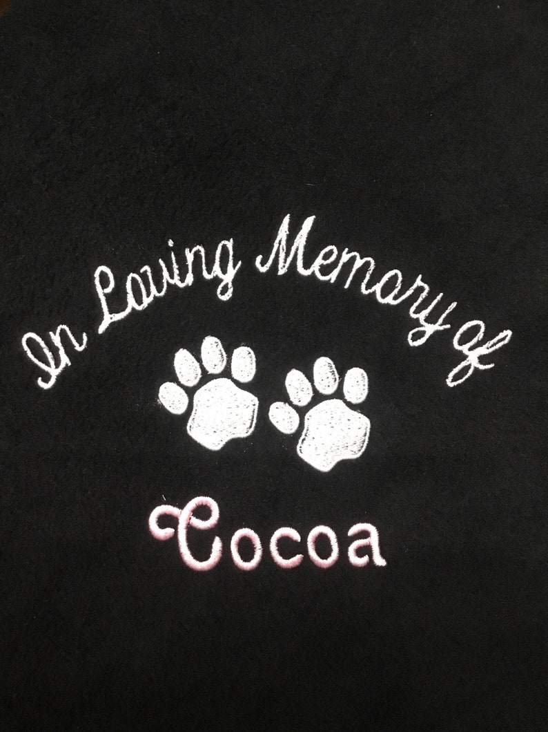 May include: Black fabric embroidered with the words "In Loving Memory of" above two white paw prints. Below the paw prints, the name "Cocoa" is embroidered in pink.