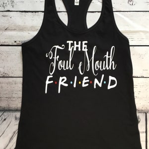 K&ouml;nnte beinhalten: Schwarzes Racerback-Tanktop mit dem wei&szlig;en Text "THE Foul Mouth FRIEND", wobei das Wort "FRIEND" im Stil der Fernsehserie Friends geschrieben ist. Das Tanktop besteht aus einem weichen Material und wird auf einer Holzoberfl&auml;che pr&auml;sentiert.