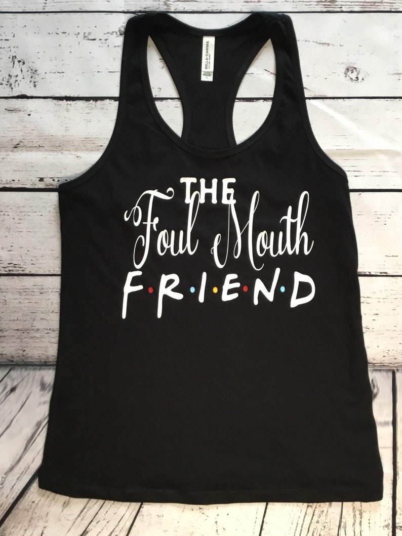 K&ouml;nnte beinhalten: Schwarzes Racerback-Tanktop mit dem wei&szlig;en Text "THE Foul Mouth FRIEND", wobei das Wort "FRIEND" im Stil der Fernsehserie Friends geschrieben ist. Das Tanktop besteht aus einem weichen Material und wird auf einer Holzoberfl&auml;che pr&auml;sentiert.