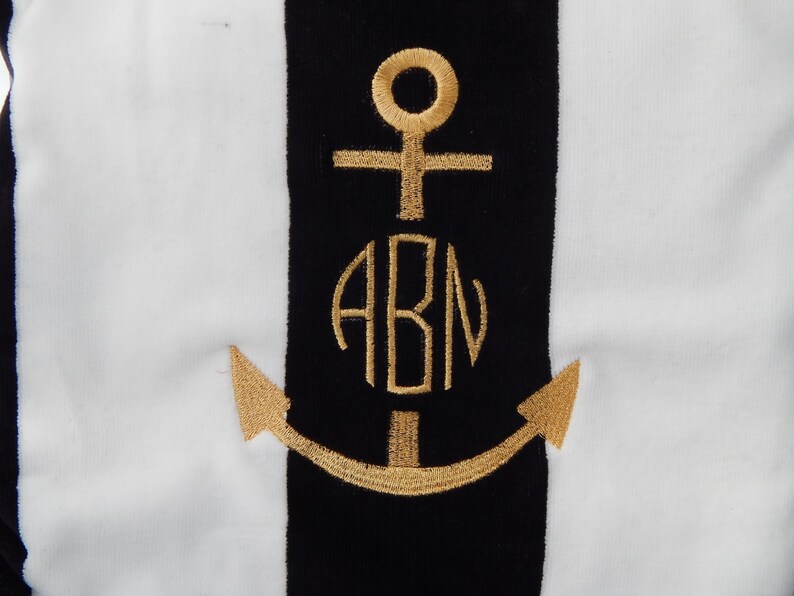 May include: A nautical-themed design featuring a gold embroidered anchor and the initials "ABN" on a black stripe. The background has vertical white stripes. The design is likely for a towel or fabric.