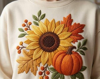 Peut inclure: Un sweat-shirt à col rond de couleur crème avec une grande broderie détaillée sur le devant. La broderie représente un tournesol jaune central, une citrouille orange, des feuilles d'érable automnales dans des tons orange et marron.