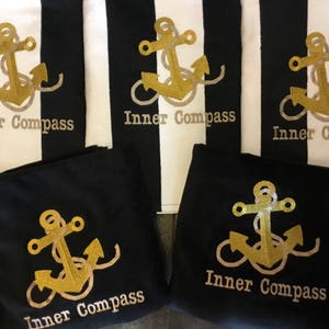 Puede incluir: Tela a rayas blancas y negras con dise&ntilde;os de anclas doradas y el texto "Inner Compass". Tambi&eacute;n, tela negra con dise&ntilde;os de anclas doradas y el texto "Inner Compass". Tema n&aacute;utico.